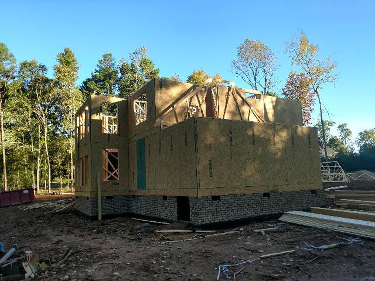 Framed house under construction with exposed wooden beams and sheathing, surrounded by mature trees and dirt ground
