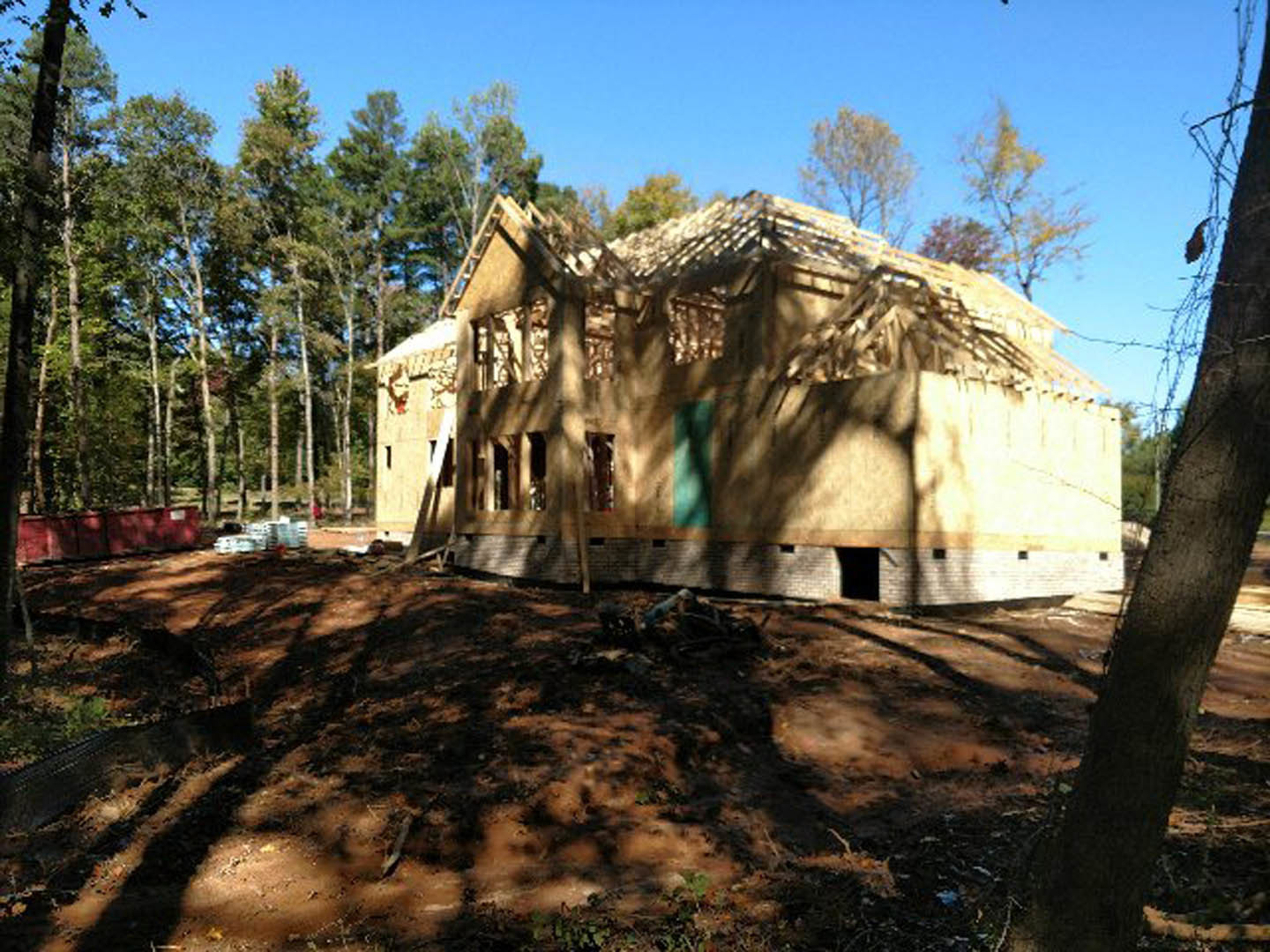 Wood-framed house under construction with exposed beams and plywood, surrounded by tall trees and dirt ground, clear sky in background