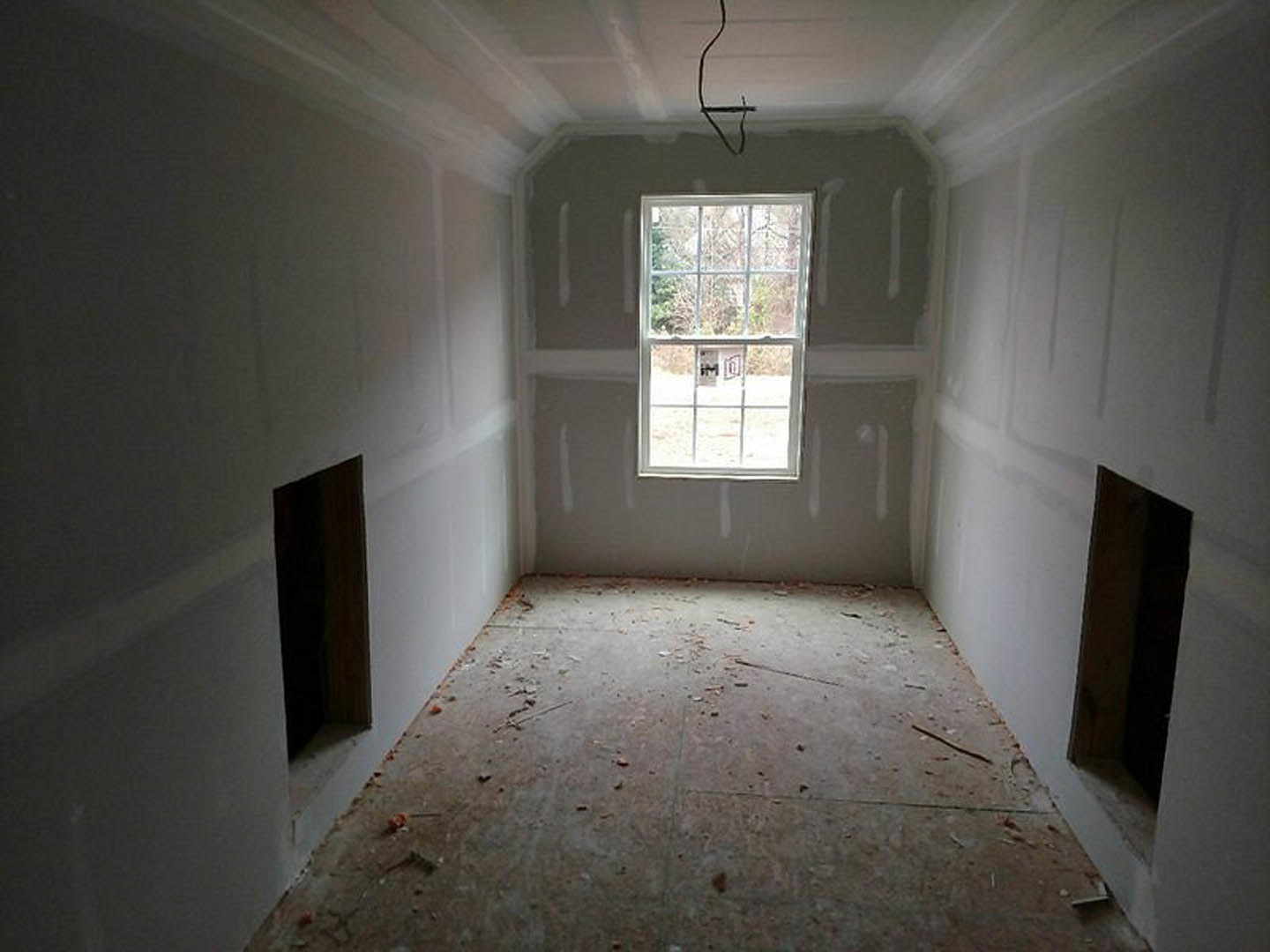 White plaster walls and large window in an unfinished room, debris scattered across the floor, black rectangular object near the window.