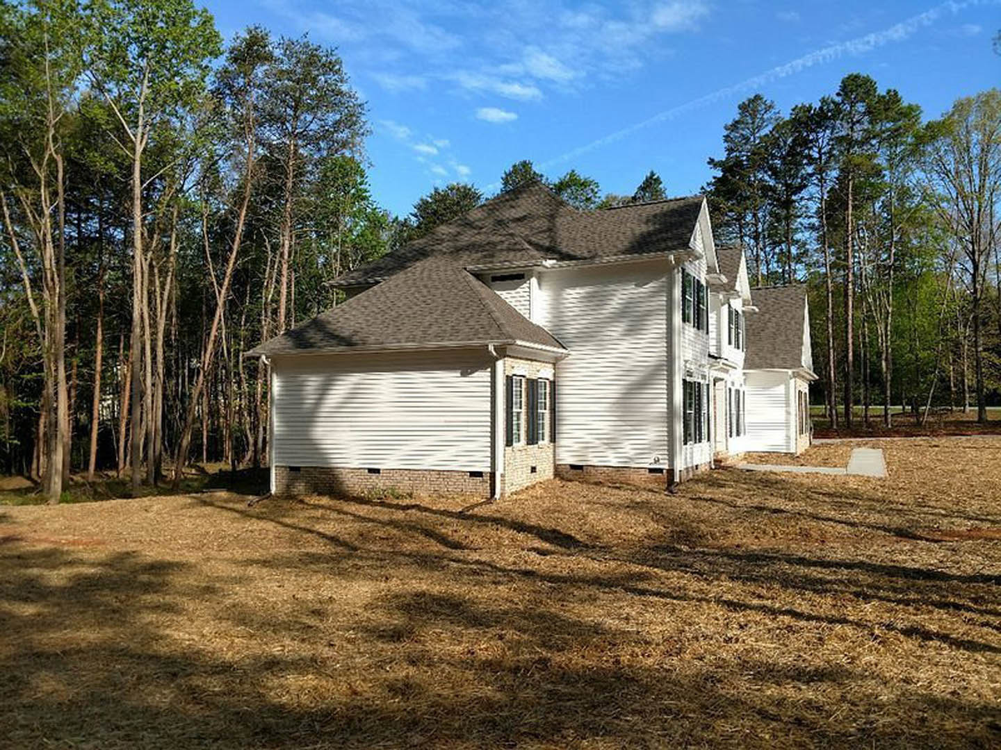 White siding house with multiple windows, patch of green grass in front, brown trim accents, trees in background, blue sky with scattered clouds overhead