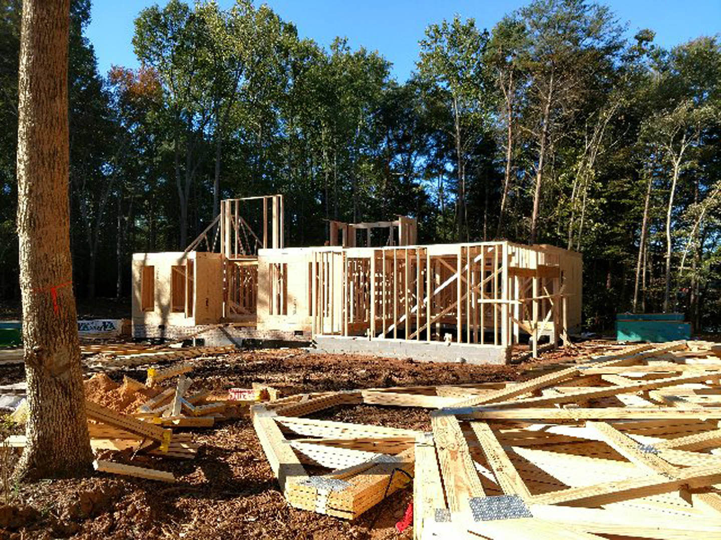 Wood-framed custom home under construction with exposed lumber, surrounded by trees and clear sky in the background
