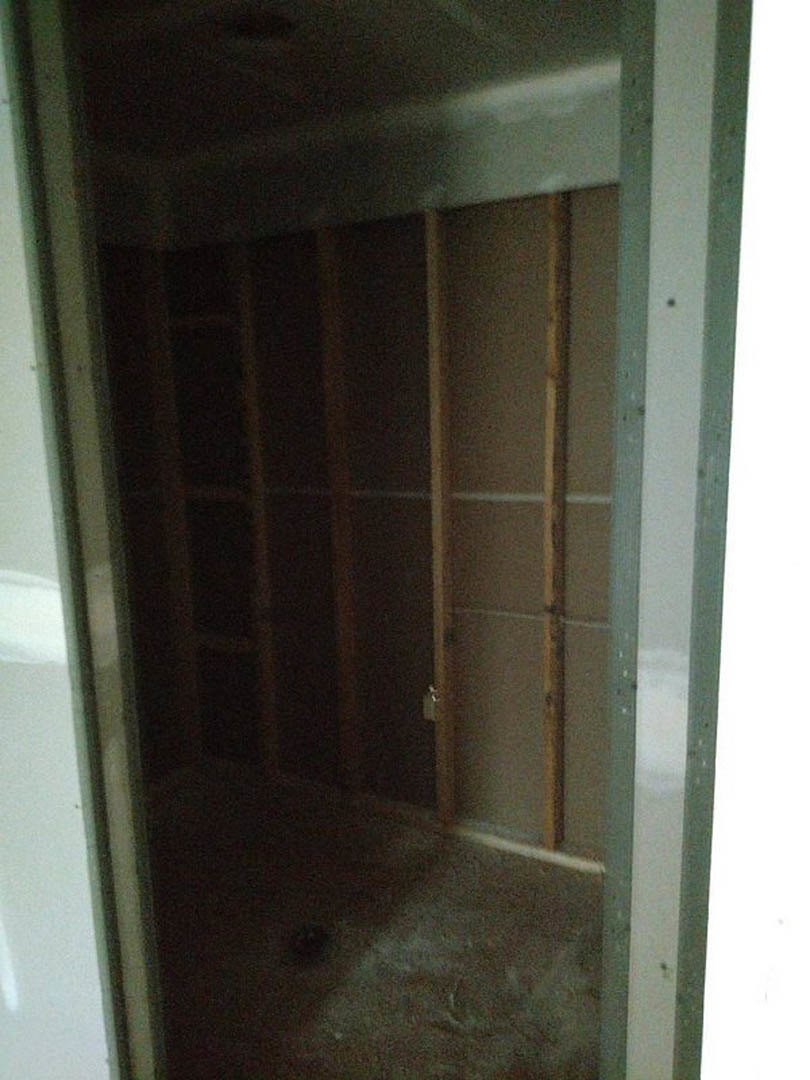 Open closet door revealing wooden shelf against white plaster wall, dark flooring, partial view of a person's head in foreground.