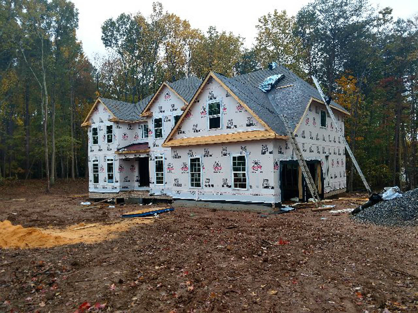 Wood-framed house under construction with exposed roof trusses, surrounded by tall trees and dirt ground, windows partially installed