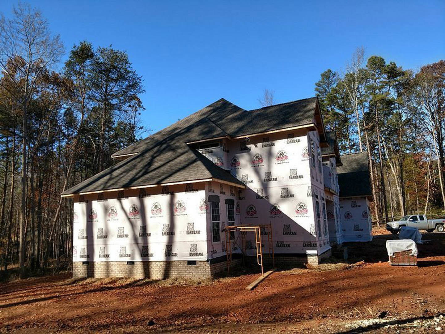 White-walled house under construction with metal ladder leaning against exterior, white truck parked nearby, dirt road and wooden ramp in foreground, trees in background.