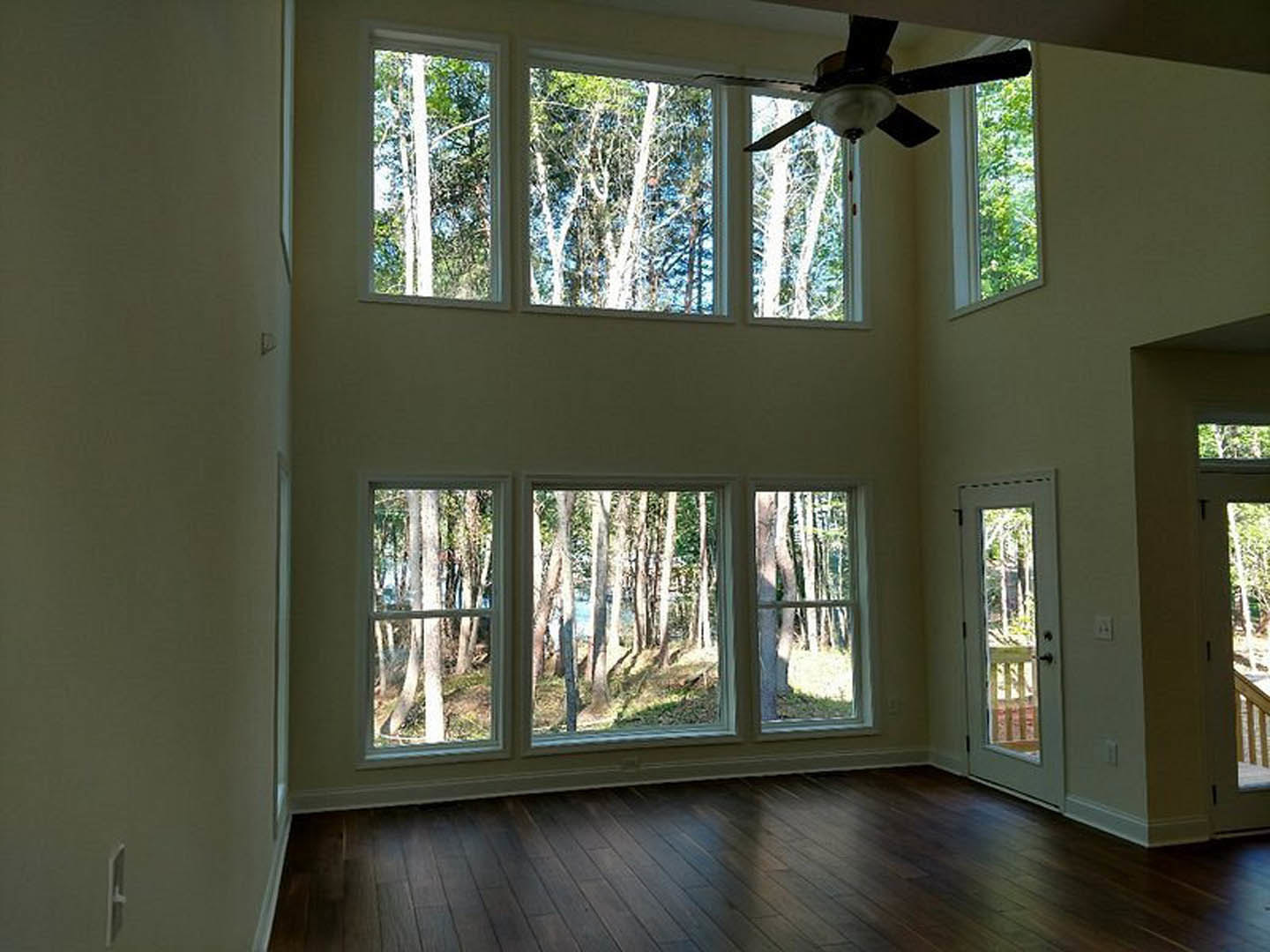 Spacious living area with multiple large windows, ceiling fan with light fixture, wooden flooring, and views of porch and trees outside