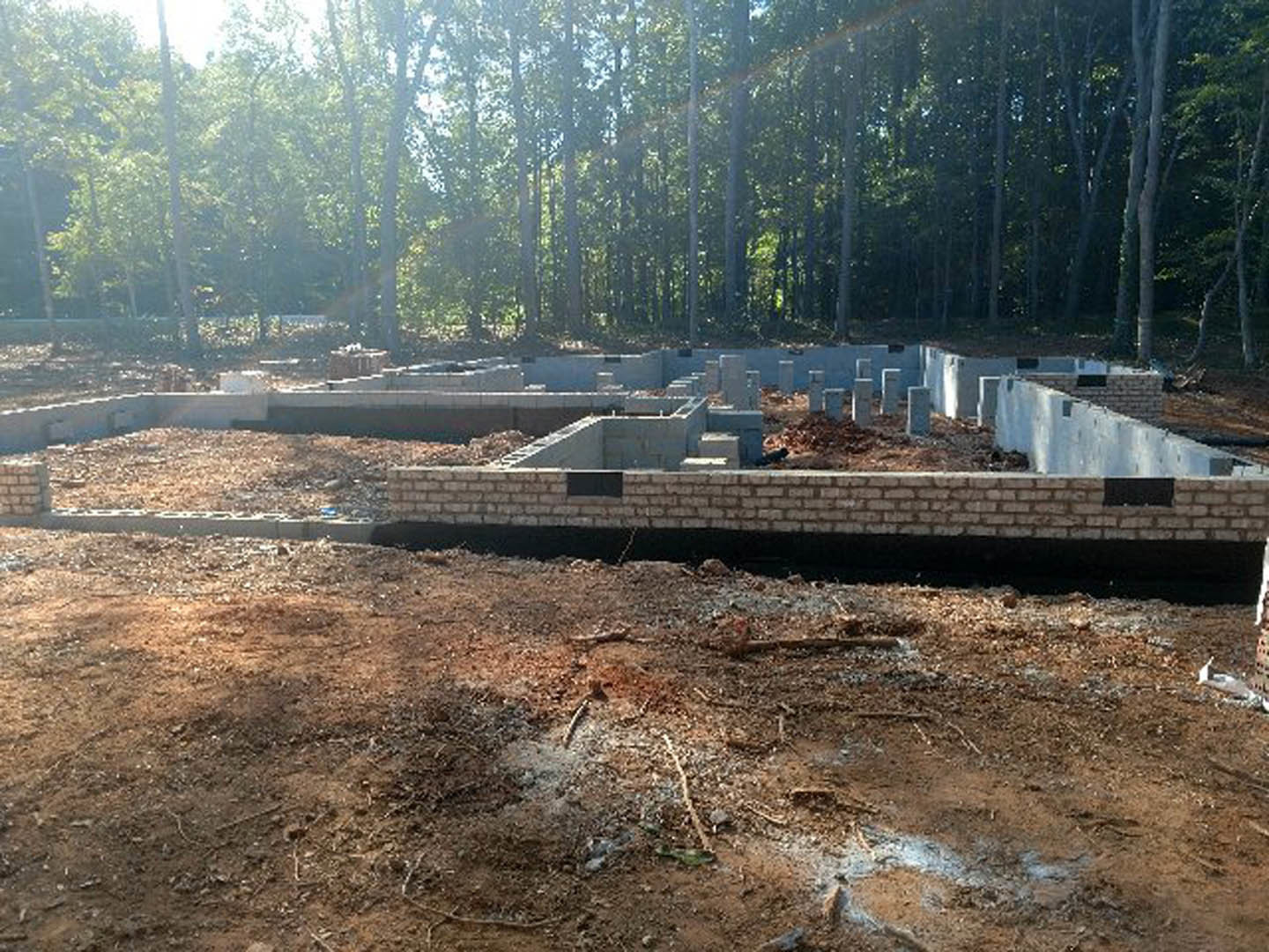 Partially built brick foundation on a dirt construction site, surrounded by mature trees and clear sky in the background