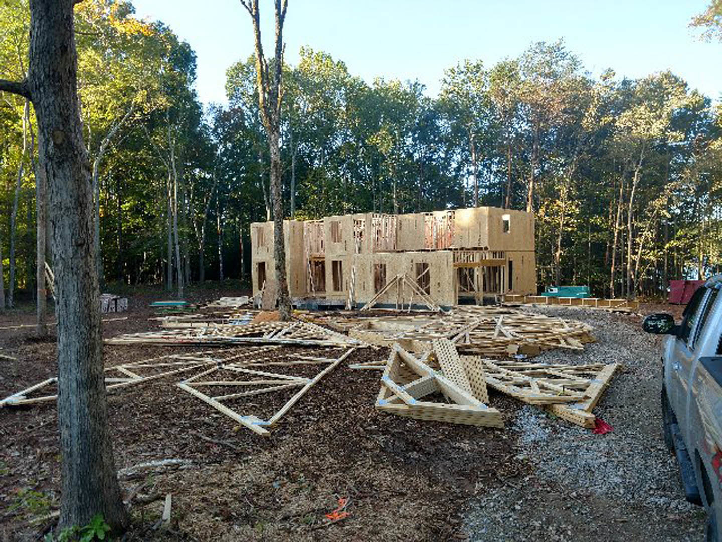 Framed custom home under construction with exposed wood beams, foundation, and surrounding trees in the background; parked car visible in foreground