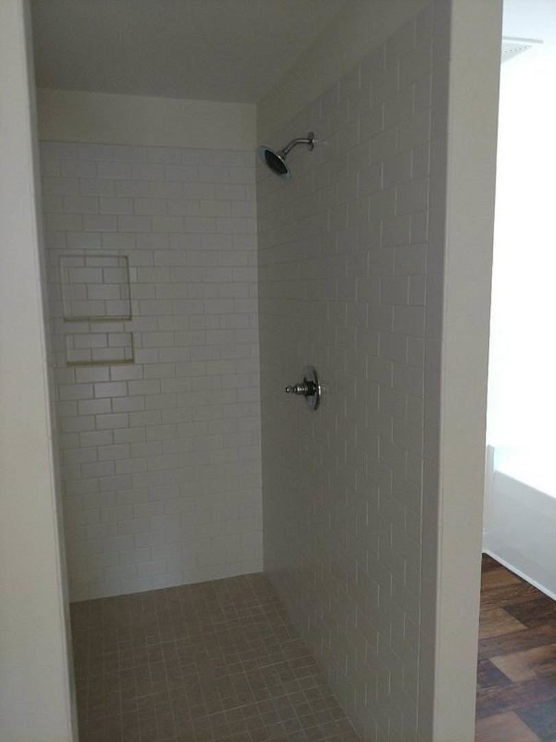 Walk-in shower with chrome shower head, white tile walls, and light wood flooring adjacent to a white door with a round metal knob.