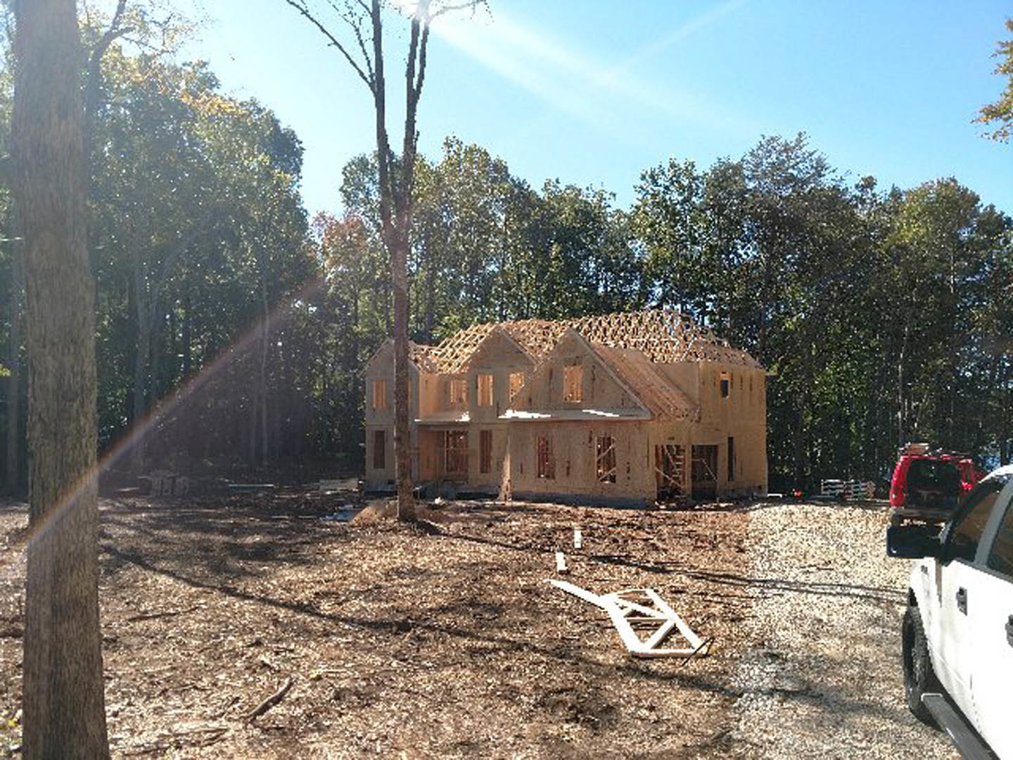 Partially built wood-framed house surrounded by tall trees, red car with open trunk parked on dirt driveway, exposed plywood walls and construction materials visible.