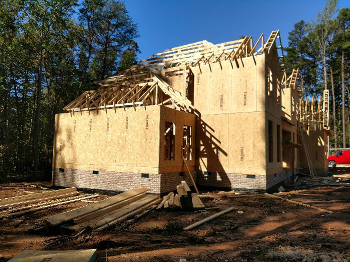 Partially built wooden house with exposed framing, roof sheathing, and construction materials scattered on dirt ground, surrounded by tall trees under a clear sky.