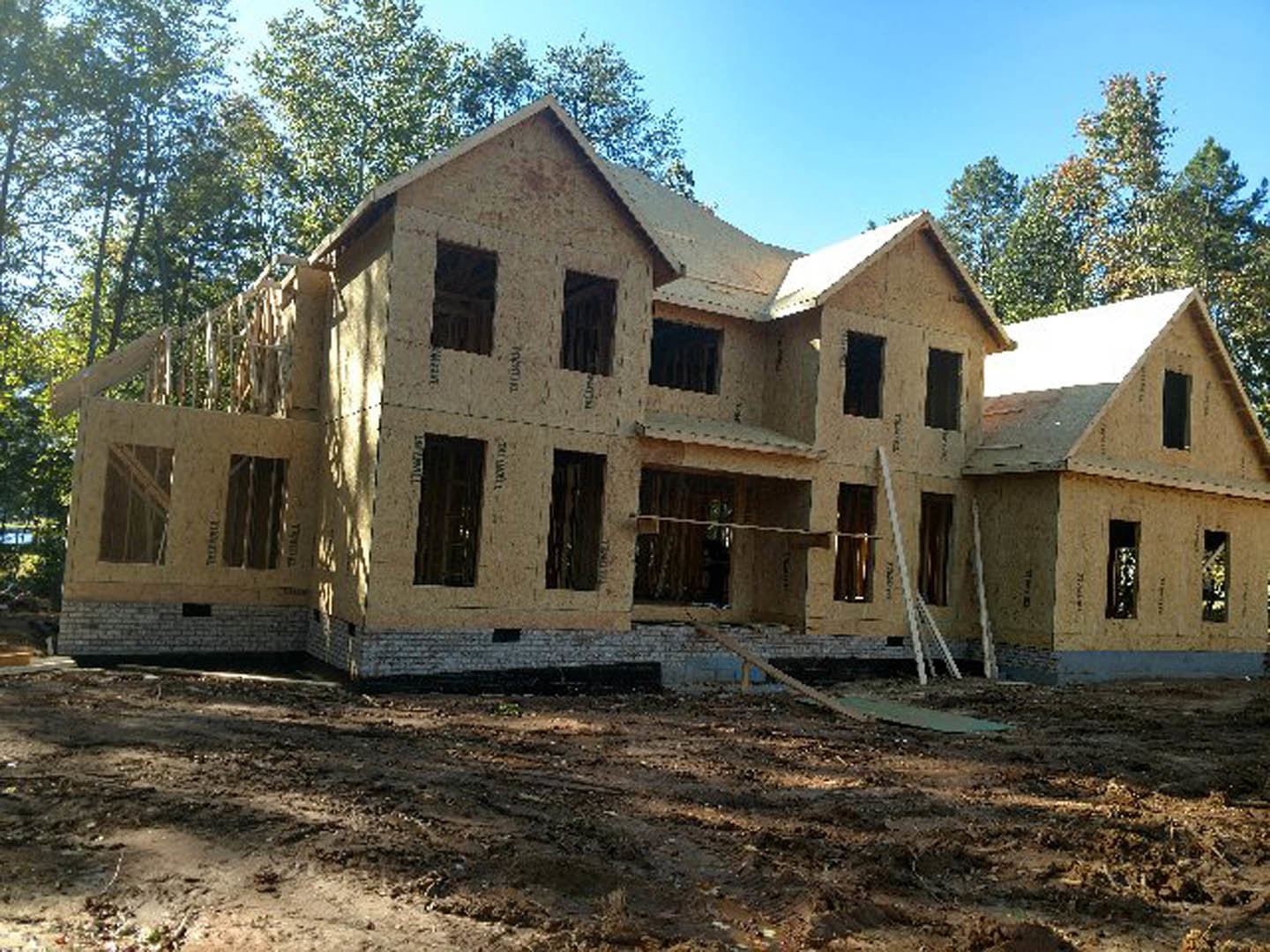 Framed house under construction with exposed lumber, partially installed windows, and surrounding mature trees