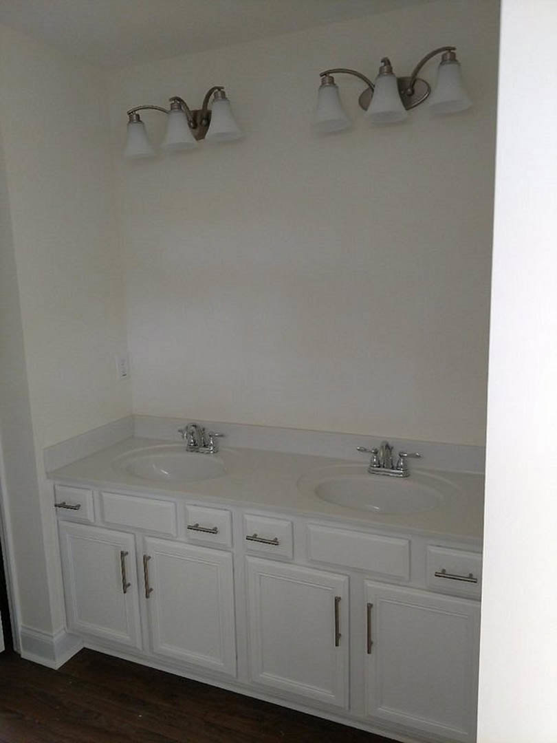 White bathroom cabinets and sinks, dark wood flooring with white trim, wall-mounted light fixture with three white shades, chrome faucet, tiled walls.