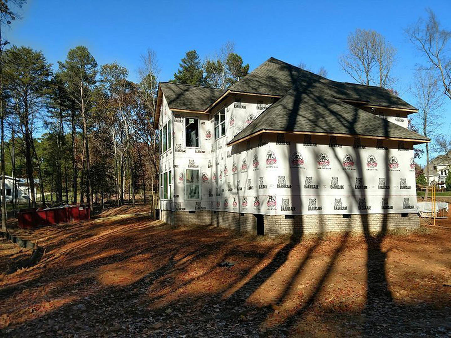 Modern house exterior with white insulated wall, large windows, tree shadows on ground, and metal-framed red container nearby