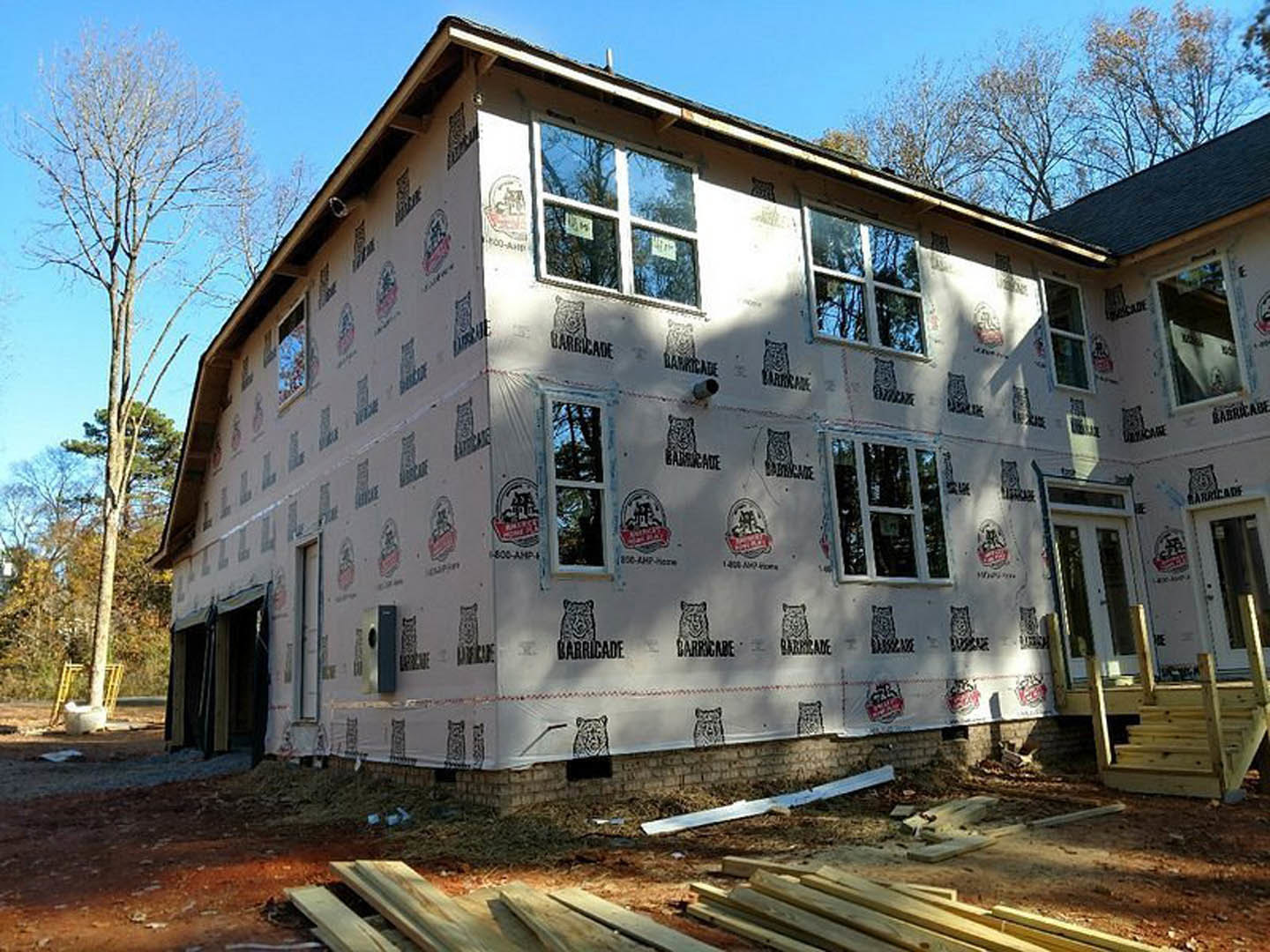 Wood-framed house under construction with exposed planks, plastic sheeting covering one wall, sash window facing leafless tree, overcast sky in background