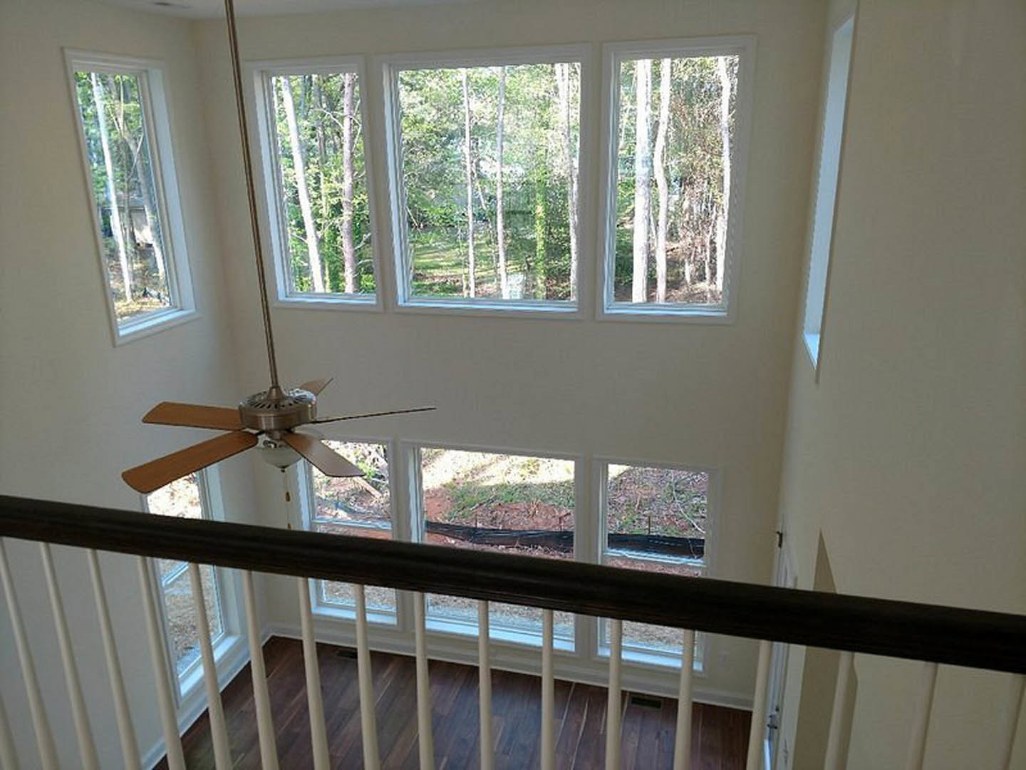 Ceiling fan with light fixture above wooden floor, large windows with trees visible outside, staircase with wooden railing and balusters.