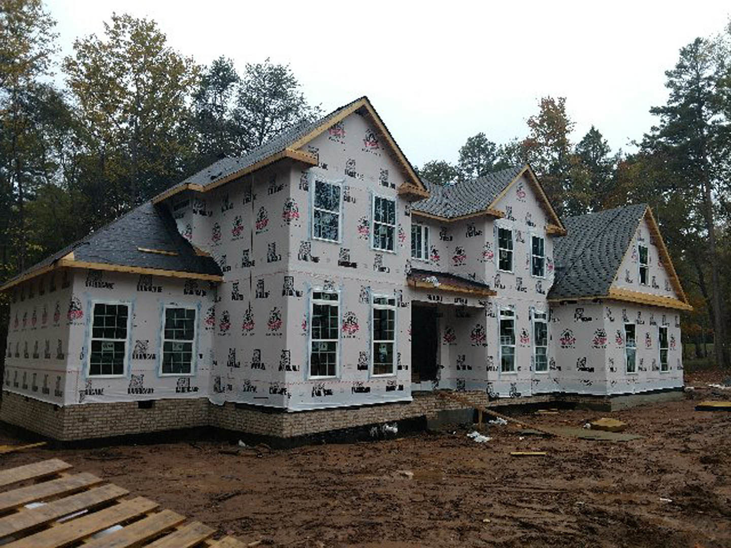 Framed house under construction with exposed plywood and roof trusses, surrounded by tall trees and clear sky