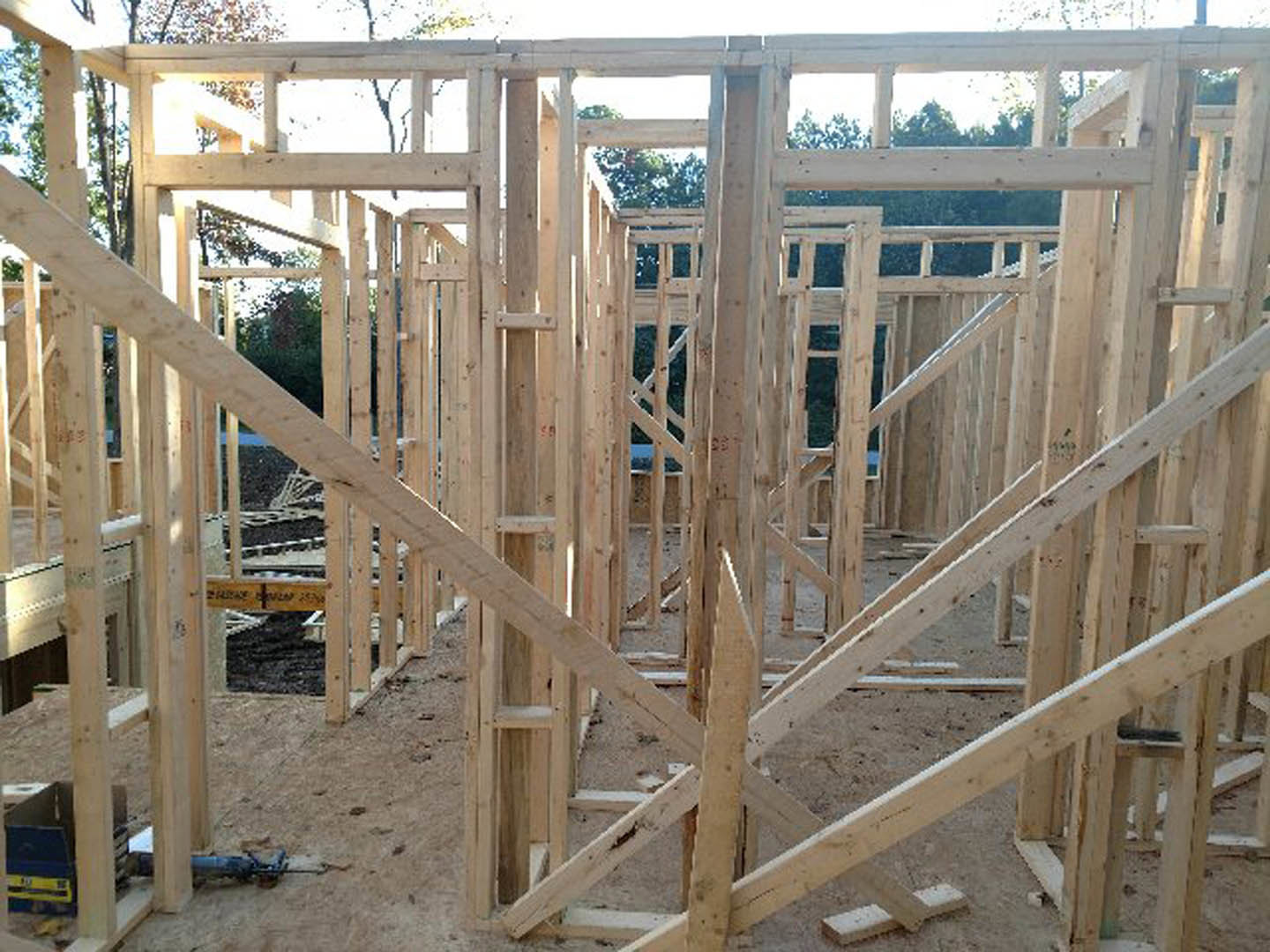 Wood-framed house under construction with exposed beams and lumber, surrounded by dirt ground and a wooden fence