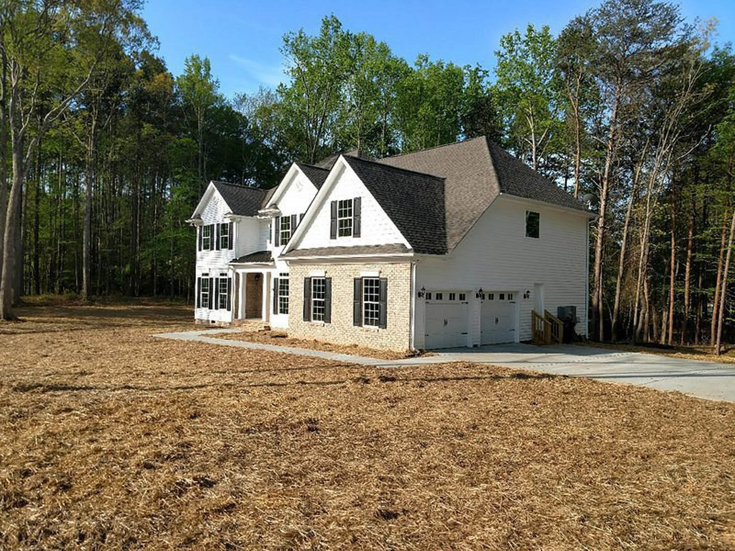 Two-story home with white siding, multi-pane windows, attached garage, wooden railing by front door, paved driveway, green lawn, mature trees in background