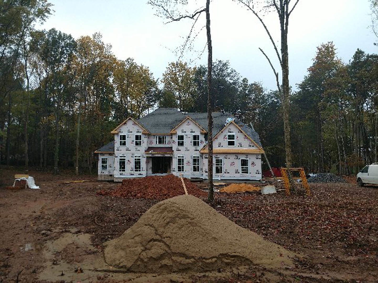 Framed house under construction surrounded by tall trees, exposed wooden beams and plywood, white van parked on dirt driveway, forested background