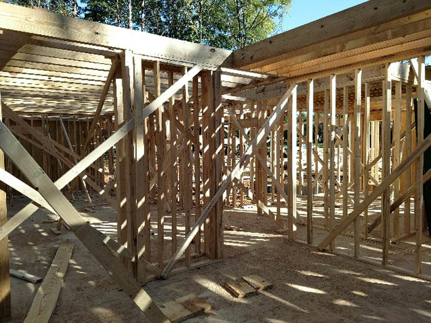 Wood-framed residential structure under construction with exposed beams, unfinished exterior walls, and stacks of lumber on dirt ground; trees and wooden fence in background.
