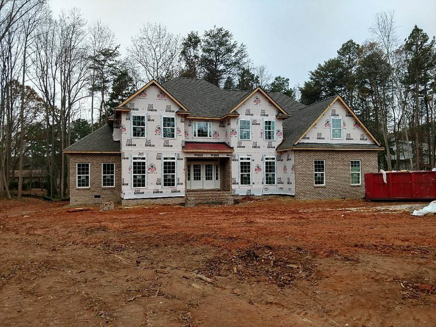 Two-story house under construction with multiple white-framed windows, exposed framing, and a red dumpster on a dirt lot with a pile of soil.