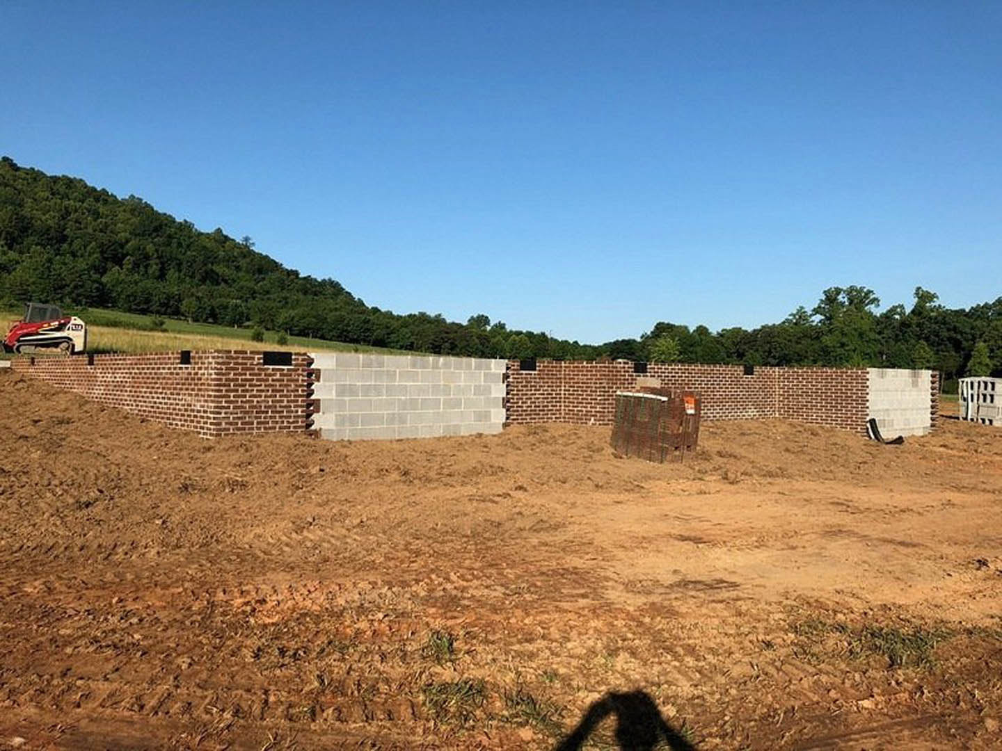 Brick wall under construction with stacked bricks and red and white tractor on dirt field, fenced boundary, grassy hill, and blue sky in background