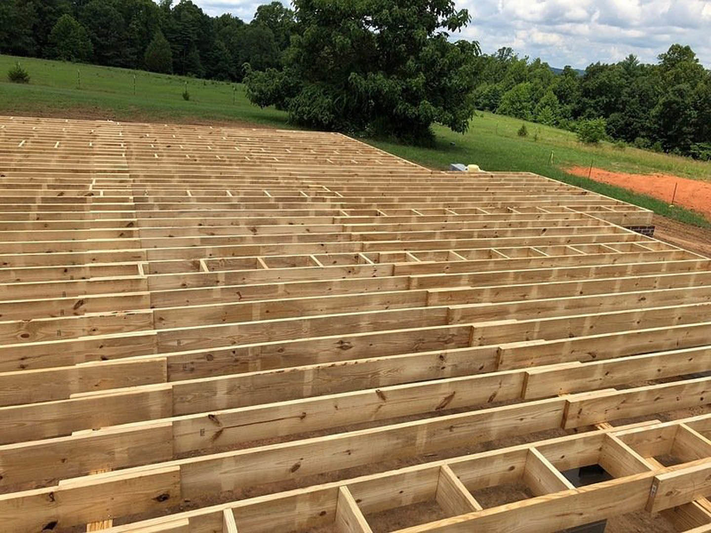 Wood-framed building under construction with exposed beams and square openings, surrounded by grass, a small bush, and a tree with green leaves; dirt path and wooden platform in