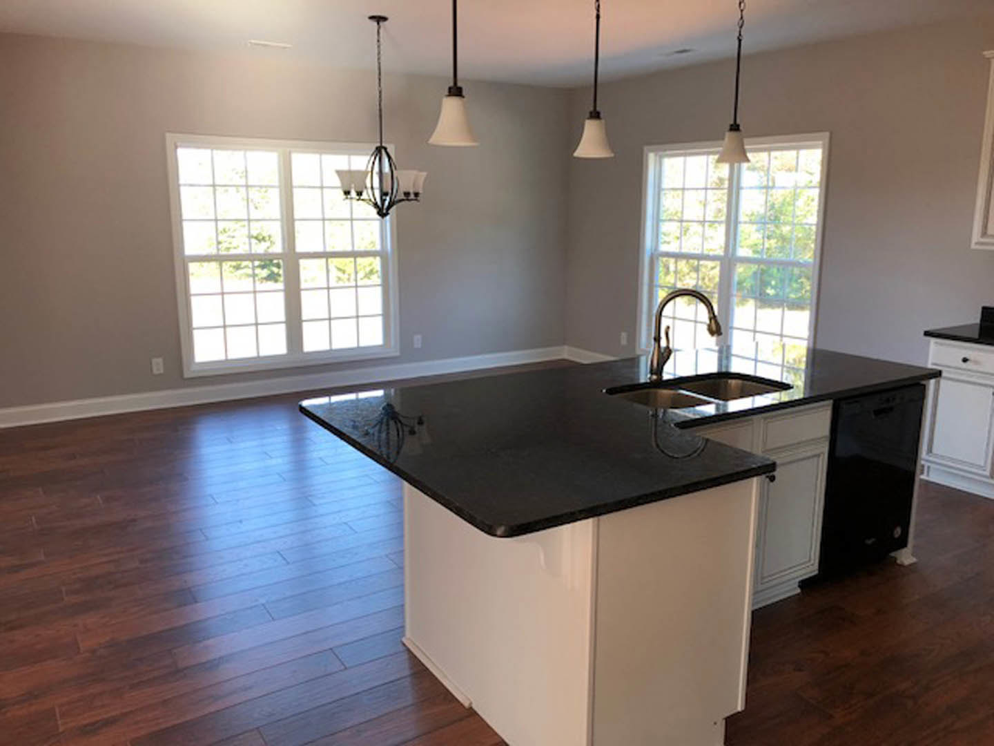Kitchen with black countertop, wood flooring, white cabinetry, stainless steel faucet and sink beneath a window, pendant lights overhead
