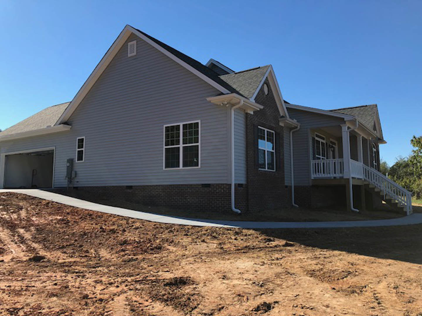 Two-story house with light siding, covered front porch with white railing, paved driveway, large windows reflecting blue sky, brick foundation, and dirt yard in foreground
