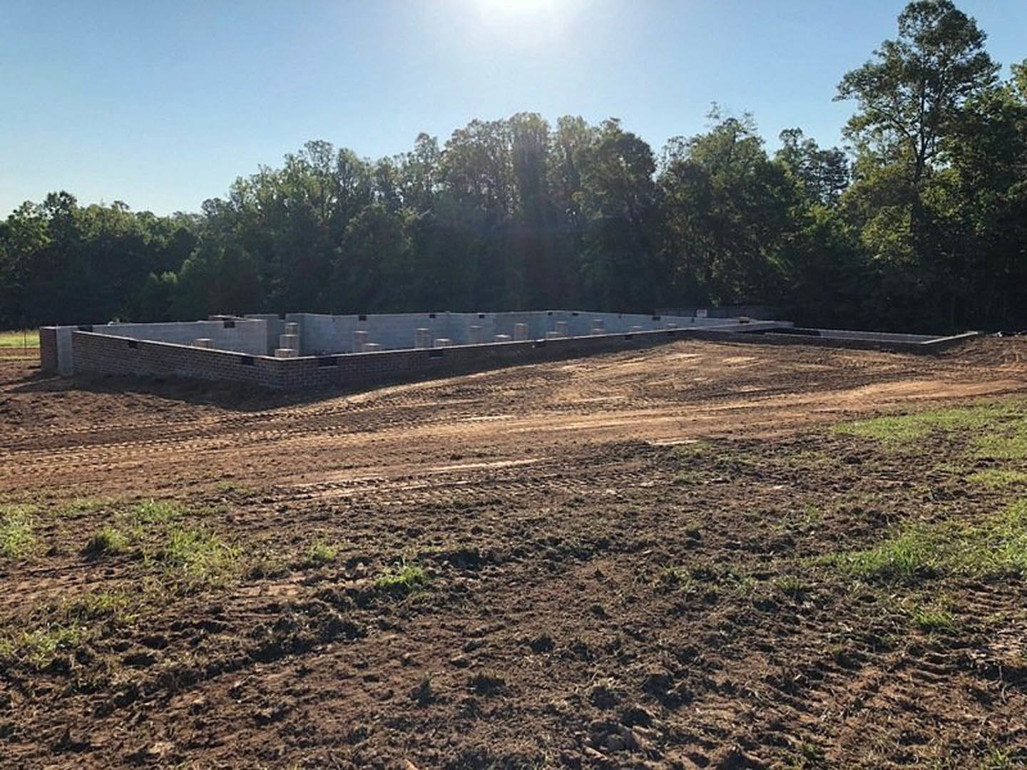 Dirt field with partially built brick wall, scattered construction materials, and leafy trees in background under clear sky