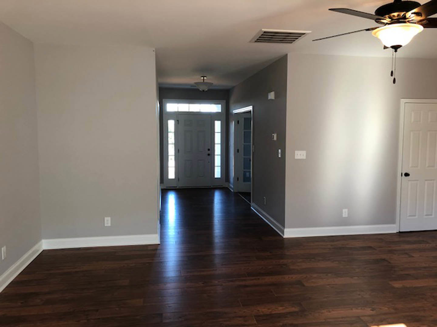 Dark wood laminate flooring with white baseboard trim, white door featuring a black handle and window, ceiling fan with integrated light fixture, wall vent visible near floor.