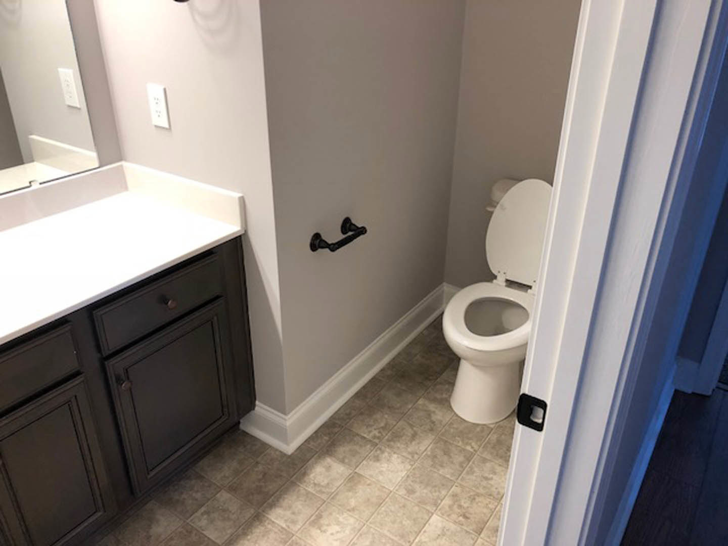 White bathroom with tile walls, modern toilet with lid up, rectangular sink set on white countertop, black drawer beneath, chrome faucet, light-colored floor tiles