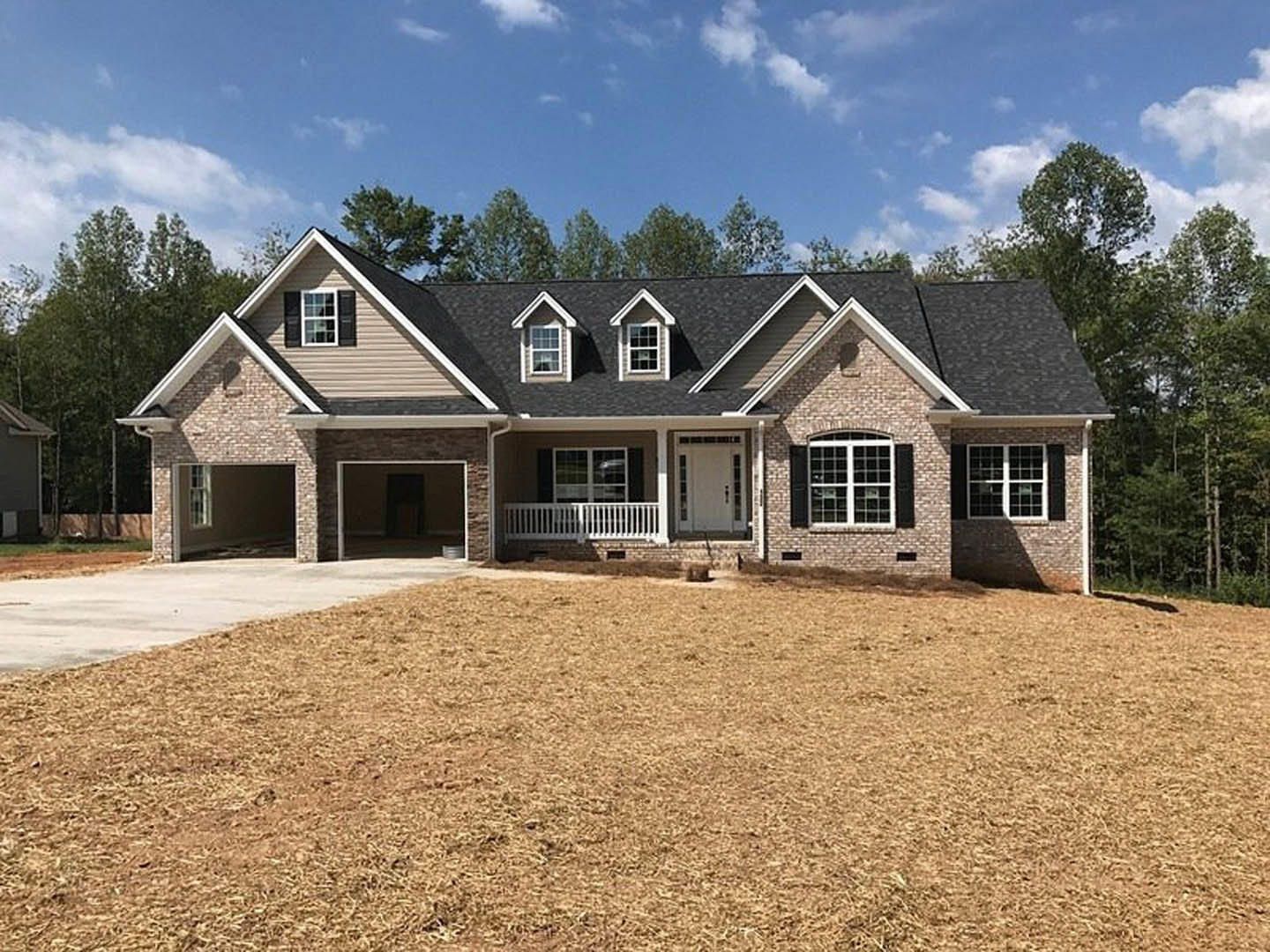 Two-story brick house with attached garage, covered front porch, multiple windows, white door with glass panes, white fence, and paved driveway bordered by grass lawn.