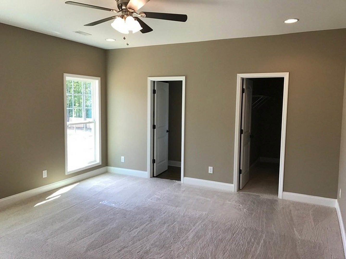 Bedroom with light wood flooring, white walls, two white doors with black hardware, ceiling fan with light fixture, window overlooking trees, open door leading to a dark adjacent
