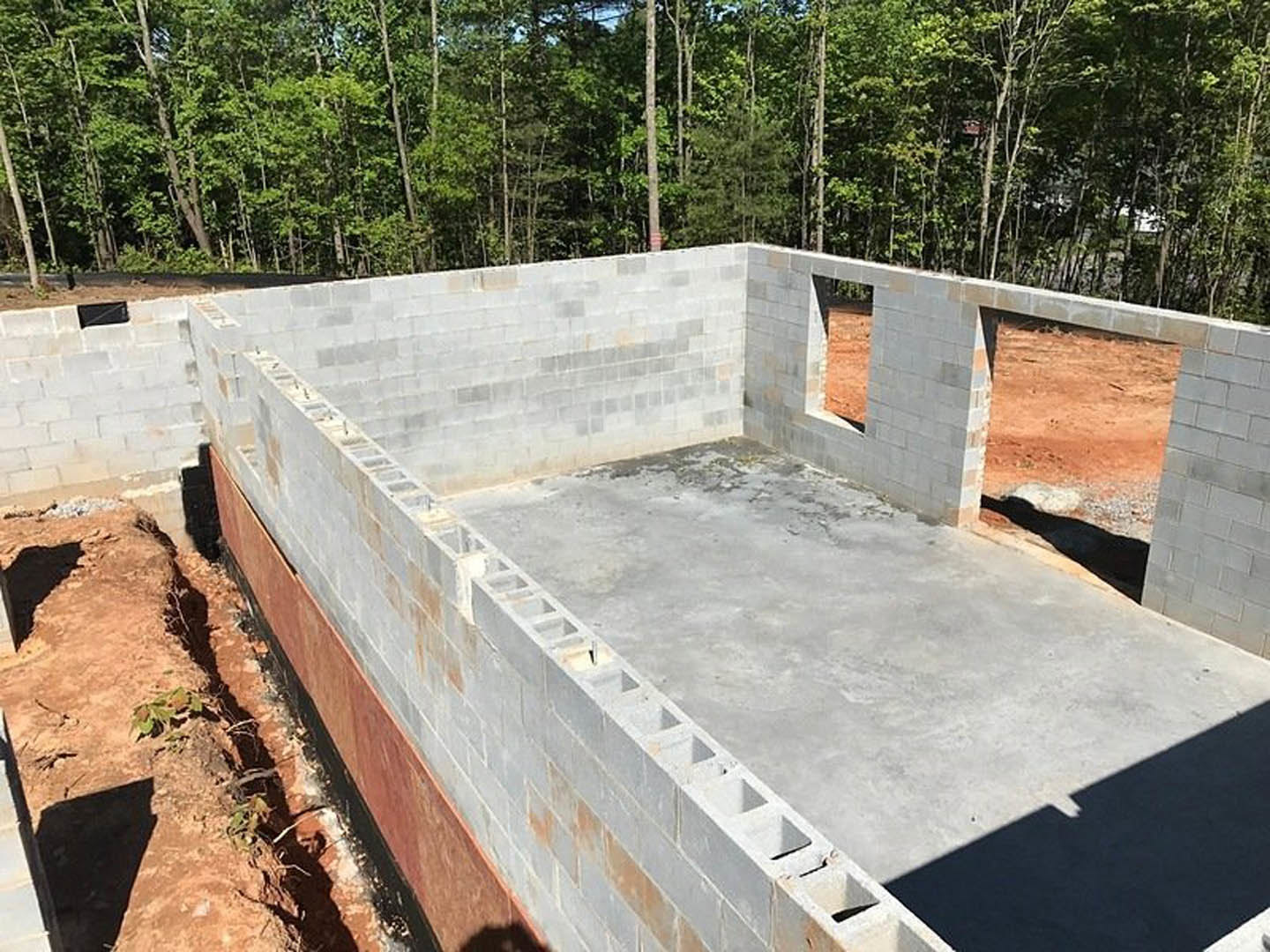 Concrete foundation with exposed hole in the ground, surrounded by dirt, plants, and tree-lined area; shadows cast on cement surface and wall.