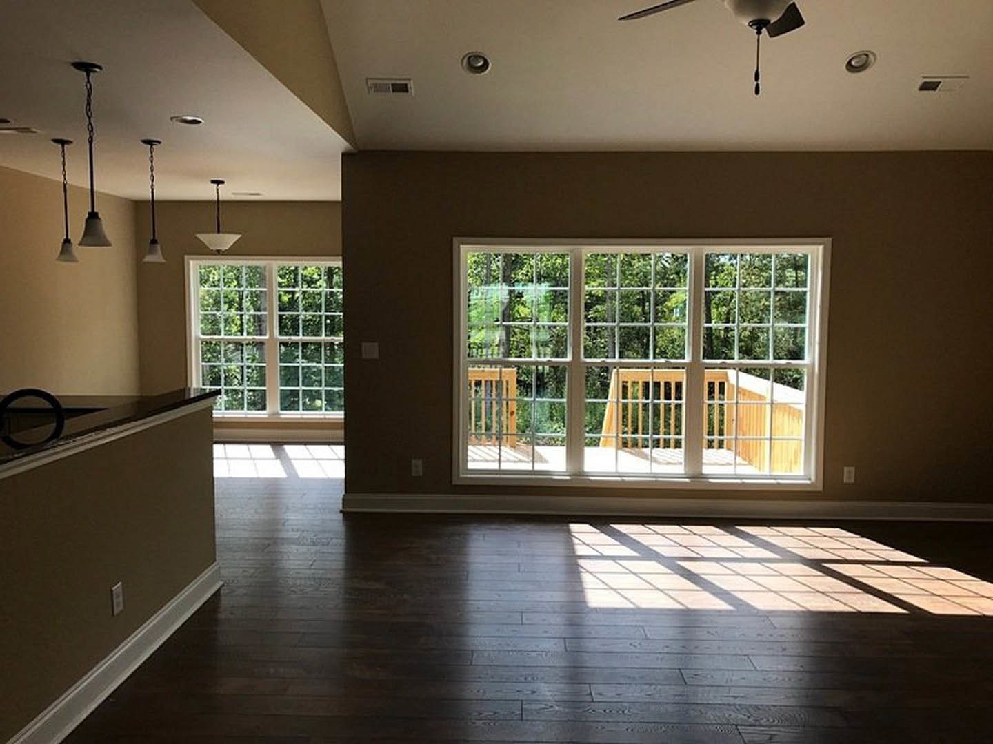 Open-concept living room with dark wood flooring, adjoining kitchen, large windows overlooking a deck and trees, ceiling lights illuminating the space