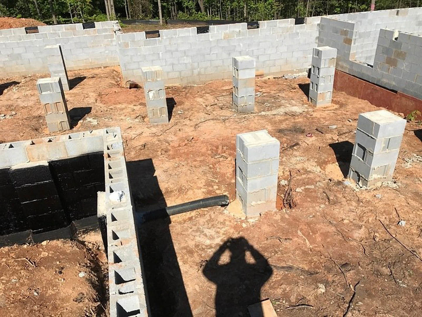 Concrete foundation with stacked black blocks, brick and concrete pillars, white utility box, construction site under sunlight with shadow of person visible on ground