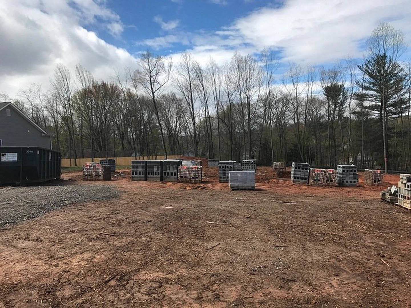 Modern farmhouse with white siding and black-framed windows, surrounded by a dirt yard, wooden fence, and scattered crates; tall trees and blue sky in the background.
