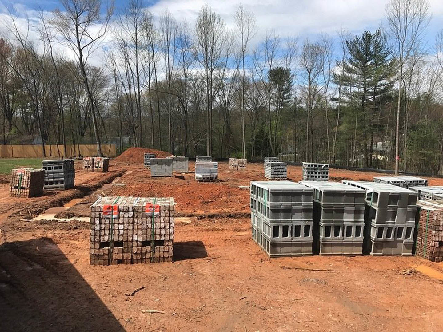 Stacks of wooden pallets and grey crates on a dirt construction site, concrete block structure with windows in background, surrounded by trees and cloudy sky