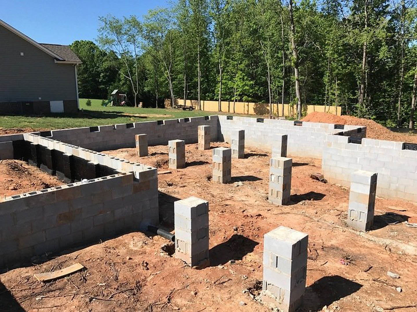 Concrete foundation with exposed pipe surrounded by soil, white brick wall partially constructed, trees in the background