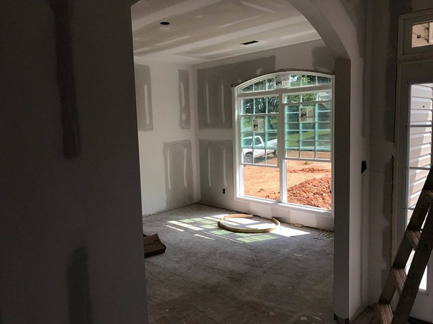 Spacious room with plaster walls, large window overlooking parking lot with white truck, brown box and white plate on floor, natural daylight illuminating interior