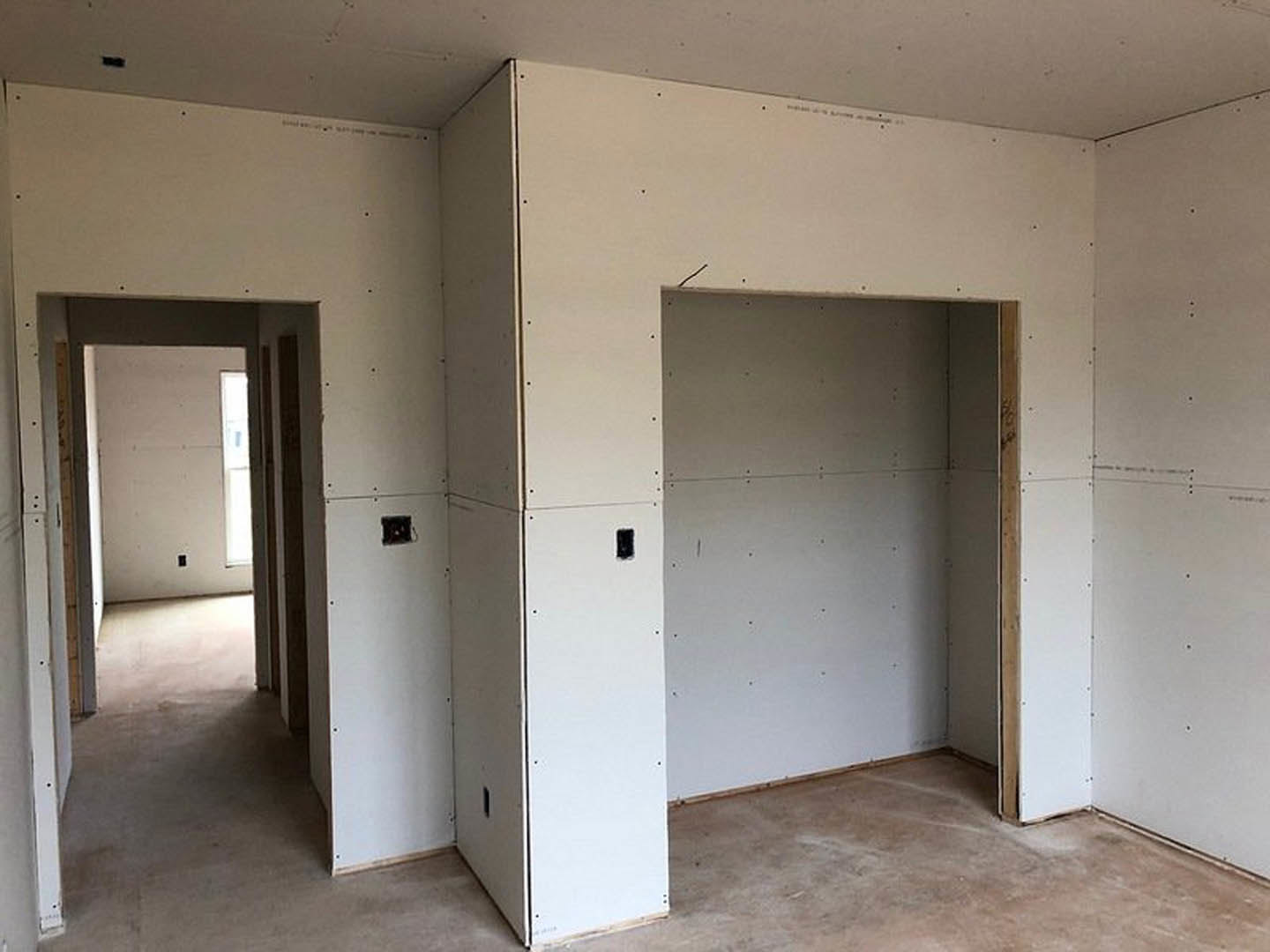 White-painted room with smooth plaster walls, black electrical outlet, doorway leading to adjacent space, light filtering through wall openings, hardwood flooring, and flat