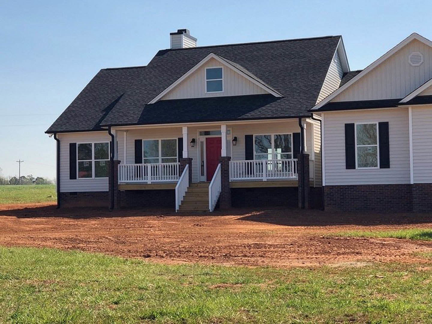 White cottage-style home with black shuttered windows, red front door, white porch railing, grassy yard, and dirt walkway leading to steps.