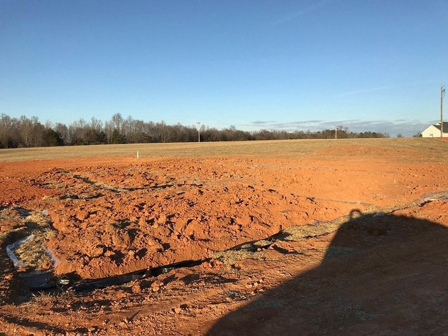 White house with black roof set on a dirt field, trees lining the background under a blue sky with scattered clouds, shadow of a person visible on dry soil in the foreground
