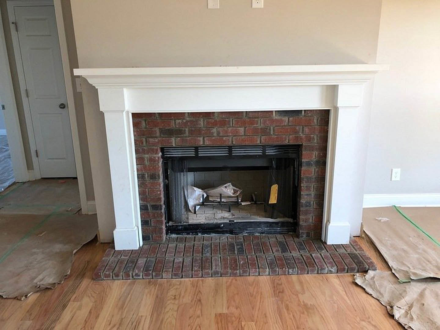 White-framed fireplace with a fish inside, adjacent to a white door with black handle, cardboard and plastic bags scattered on wood flooring