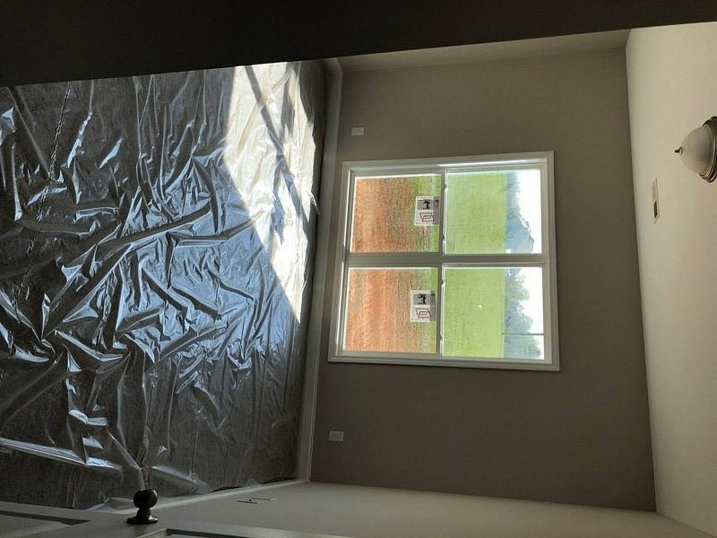 Unfinished room with white plaster walls, plastic sheeting covering surfaces, and a window overlooking a grassy field.