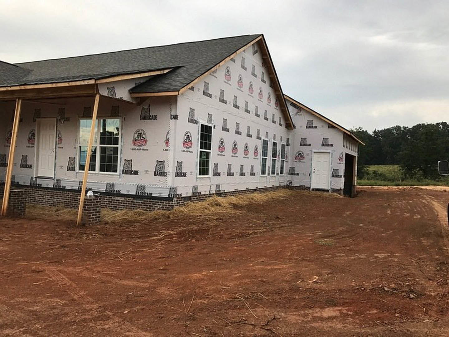 Partially built house with exposed framing, white window frames, white door with black knobs, sticker on siding, dirt and hay covering the ground, cloudy sky overhead