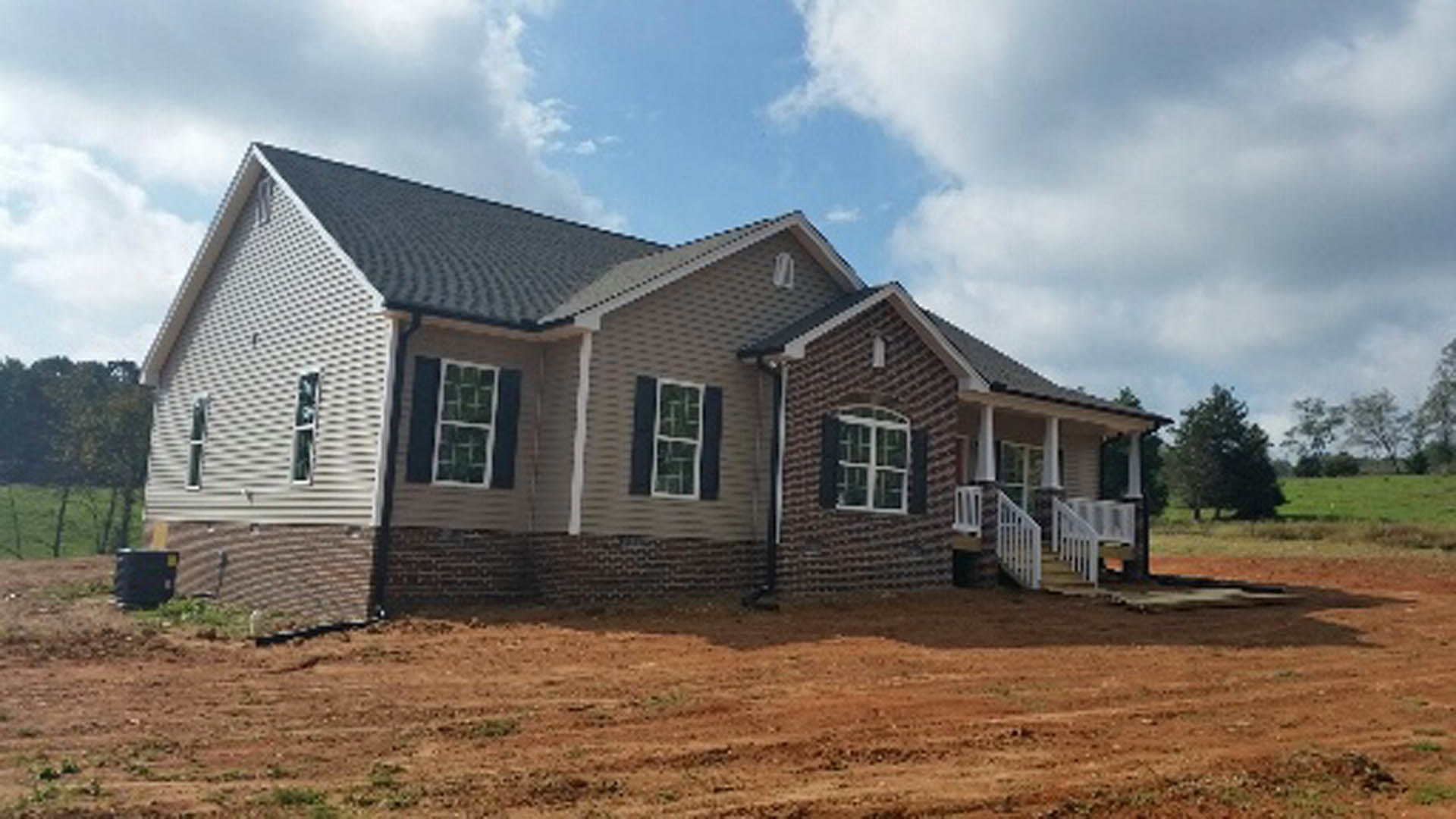 Two-story house with white siding and brick accents, surrounded by a dirt yard, blue sky overhead, white-framed windows, and a black container with yellow label near the entrance