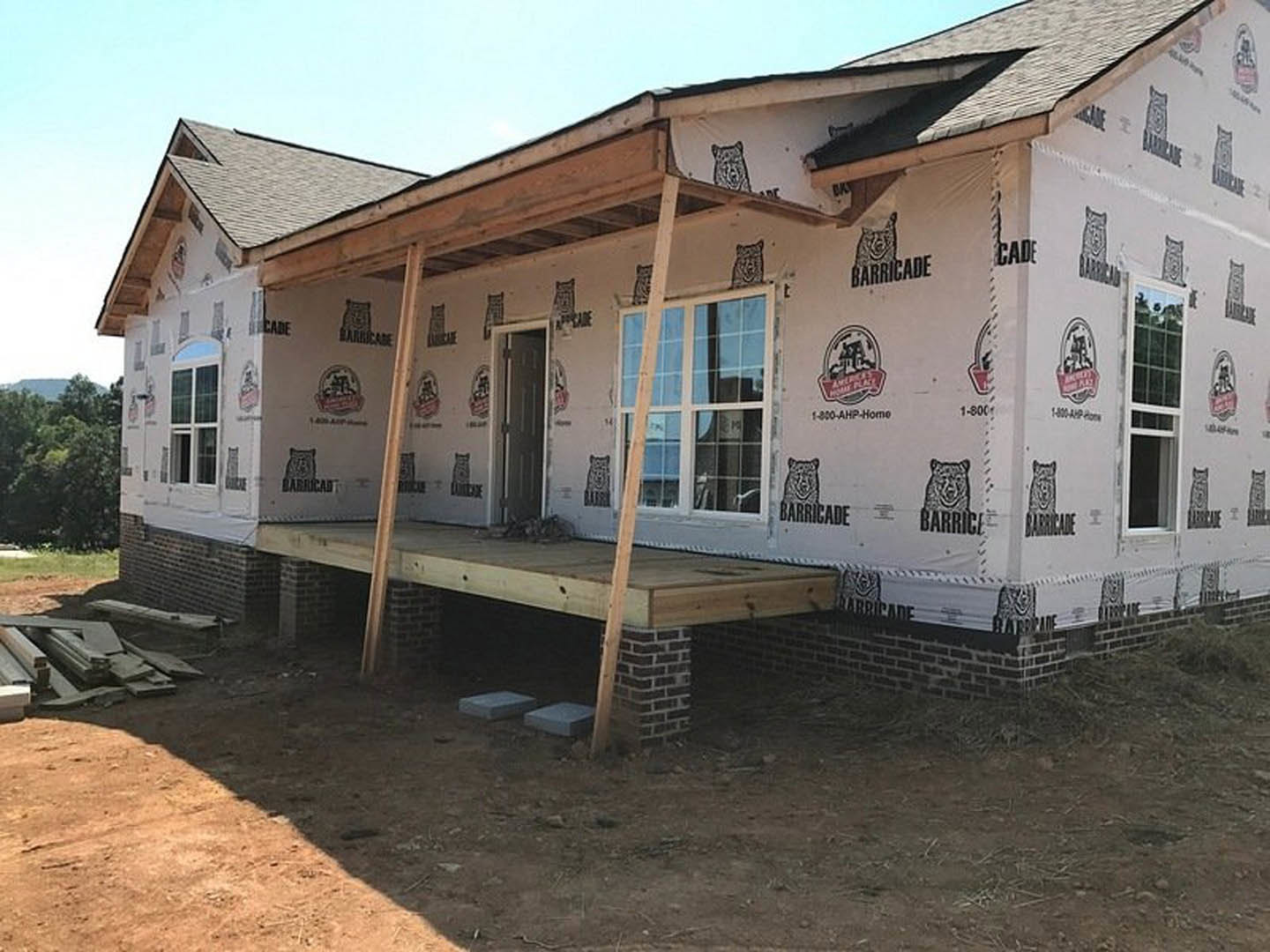 Framed house under construction featuring a covered porch with exposed wooden beams, unfinished exterior walls, and visible window openings