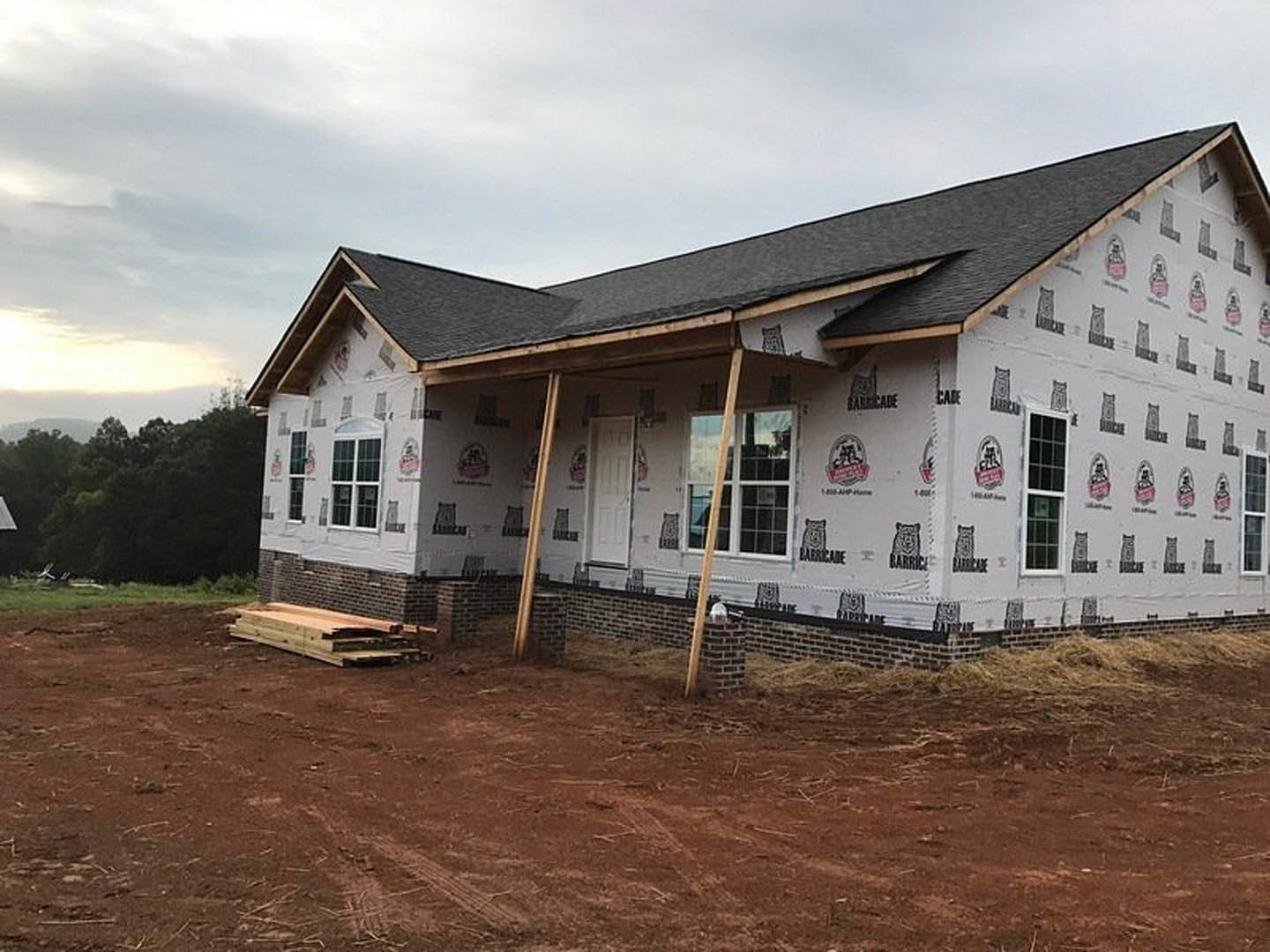 Partially built house with covered porch, exposed framing, stack of lumber on dirt yard, brick retaining wall, forested background, logo sign on exterior wall
